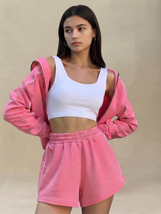 A woman modeling a pink sweatshirt and matching shorts set, worn open to reveal a white crop top, against a plain beige background.